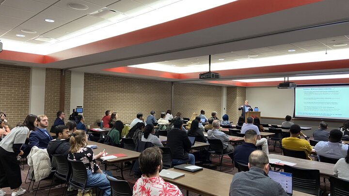 Audience seated in a conference room, listening to a presenter near a large screen displaying text.